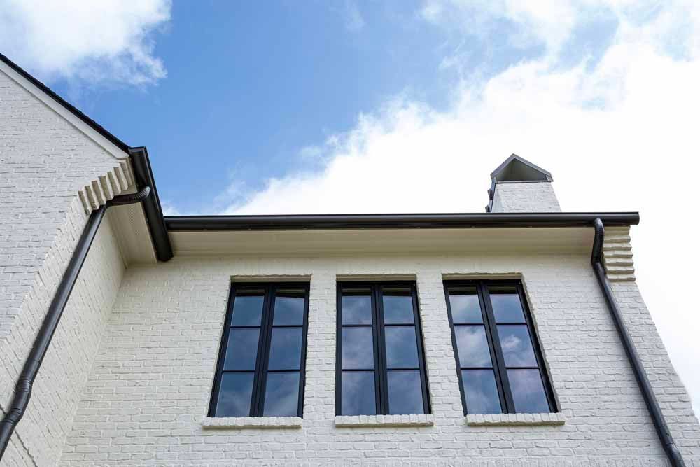Low-angle view of a white brick building featuring three dark-framed windows against a blue sky with scattered clouds.