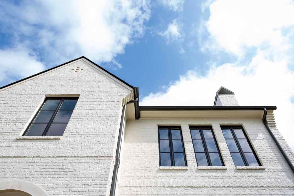 A low-angle view of a white brick building with dark-framed windows under a bright blue sky with scattered clouds.