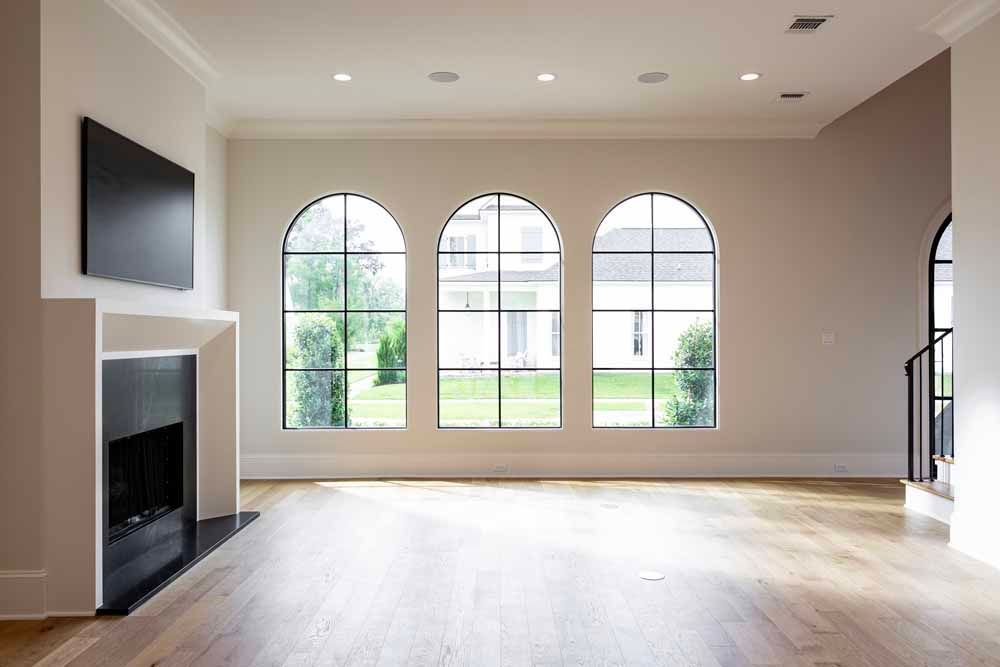 Modern living room with light floors, a black fireplace and television, and three large, arched, black-framed windows.
