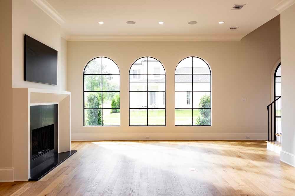 A modern living room with light wood floors, a fireplace with a wall-mounted television, and three arched windows.