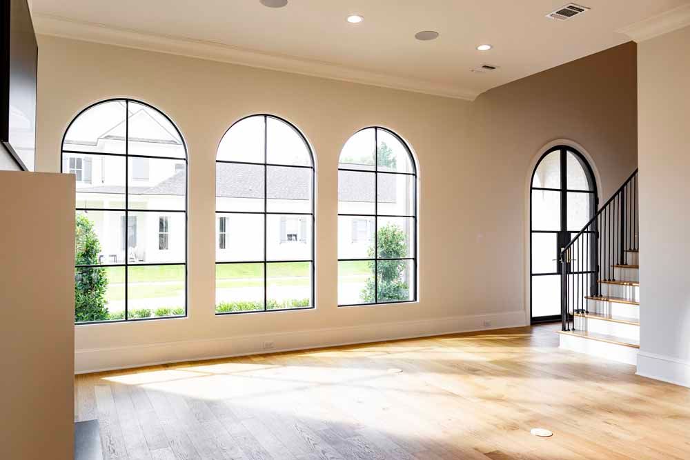 A brightly lit room with light wood floors, three tall arched windows, and a staircase with a black metal railing.