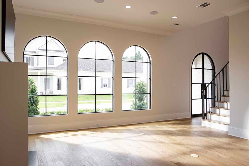 A sunlit room with three large arched windows, hardwood floors, and an arched doorway leading to a staircase.