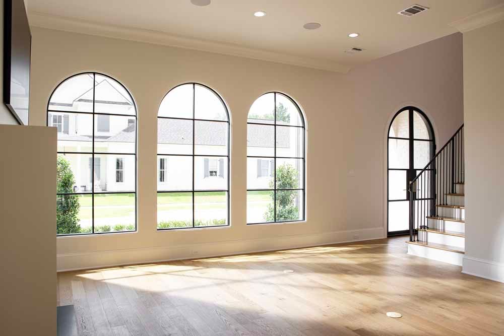 Spacious sunlit room with three large arched windows, light wood floors, and an arched doorway leading to a staircase.