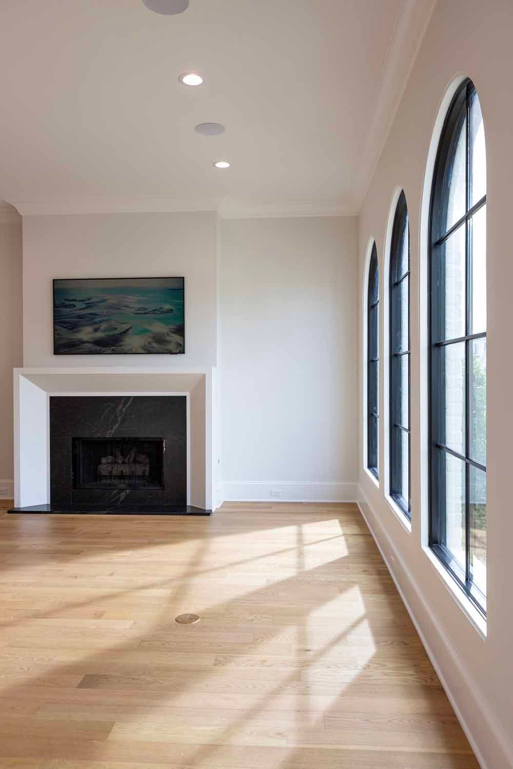 A bright, minimalist room with light wood floors, a black fireplace, and three arched windows with black frames.