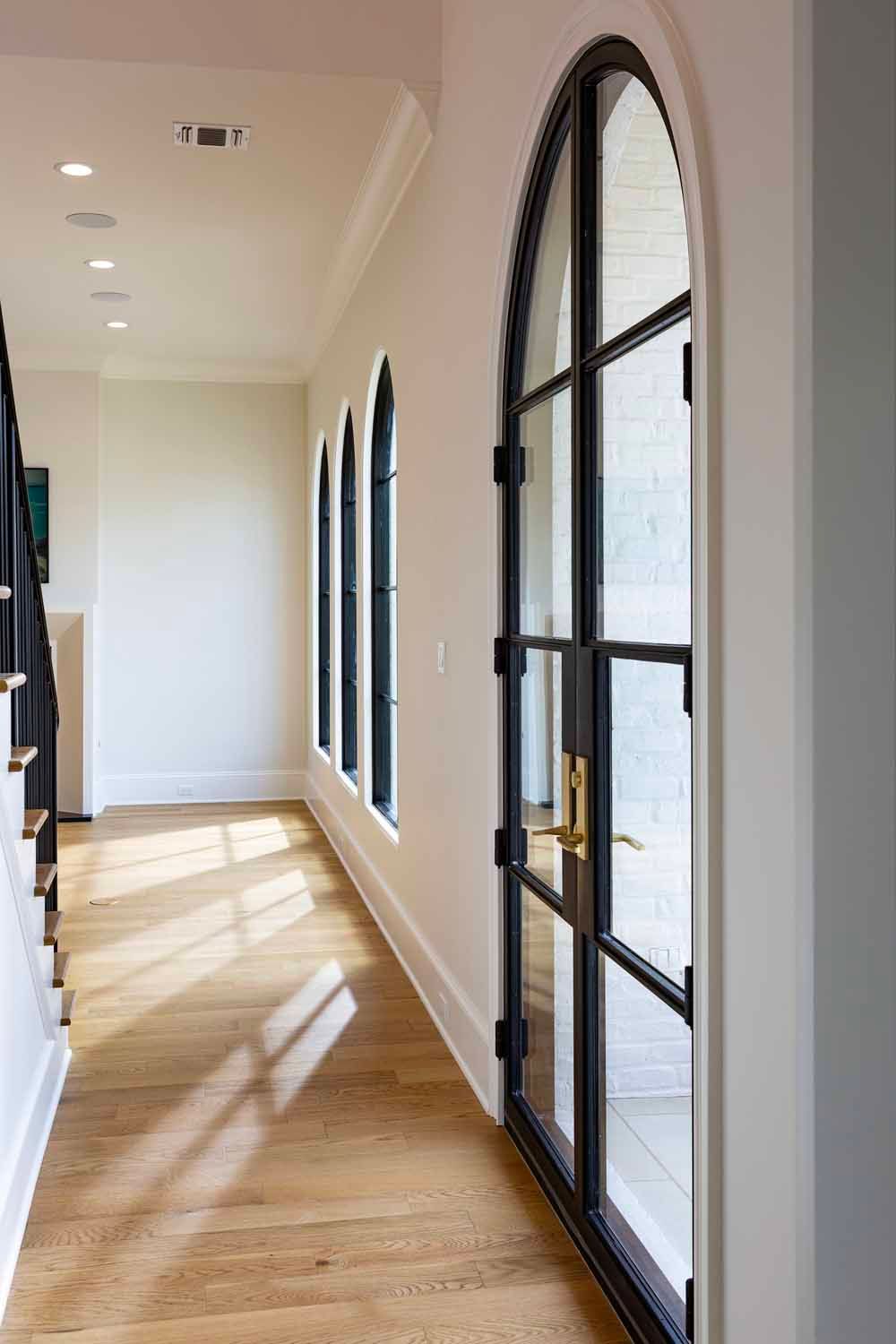 A hallway with light wood floors and white walls, featuring a series of arched, black-framed glass doors and windows.