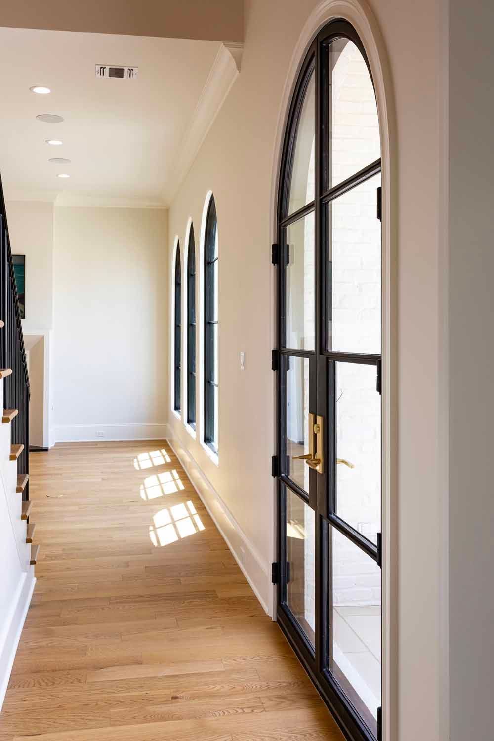 A bright hallway with light wood floors, white walls, and a row of black-framed arched windows and doors.