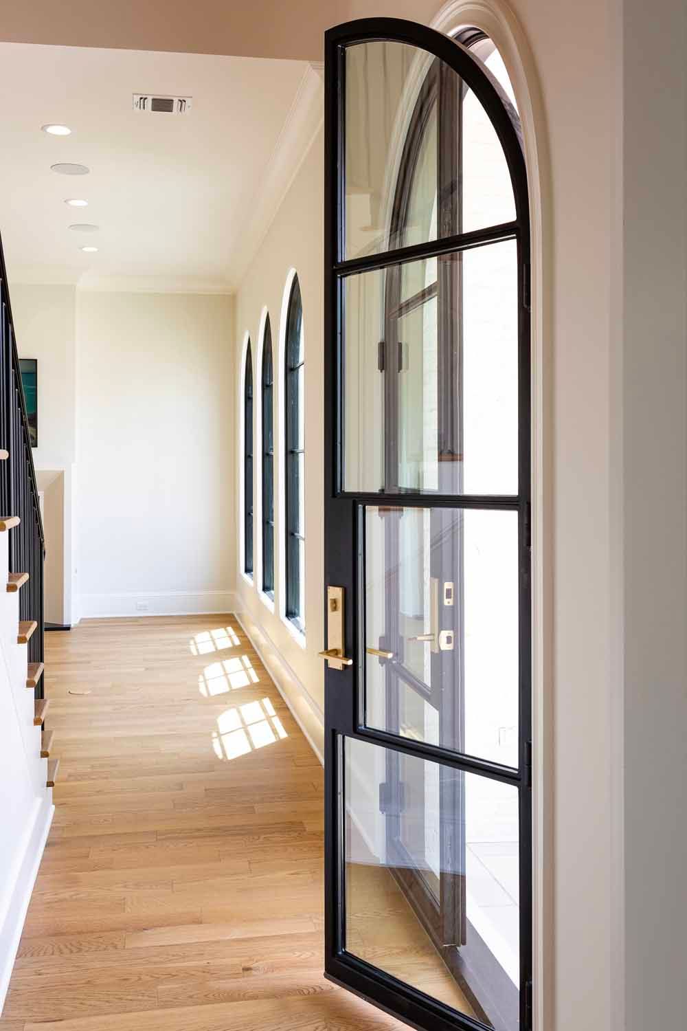 A hallway with wooden floors and an open arched black-framed glass door with a gold handle.