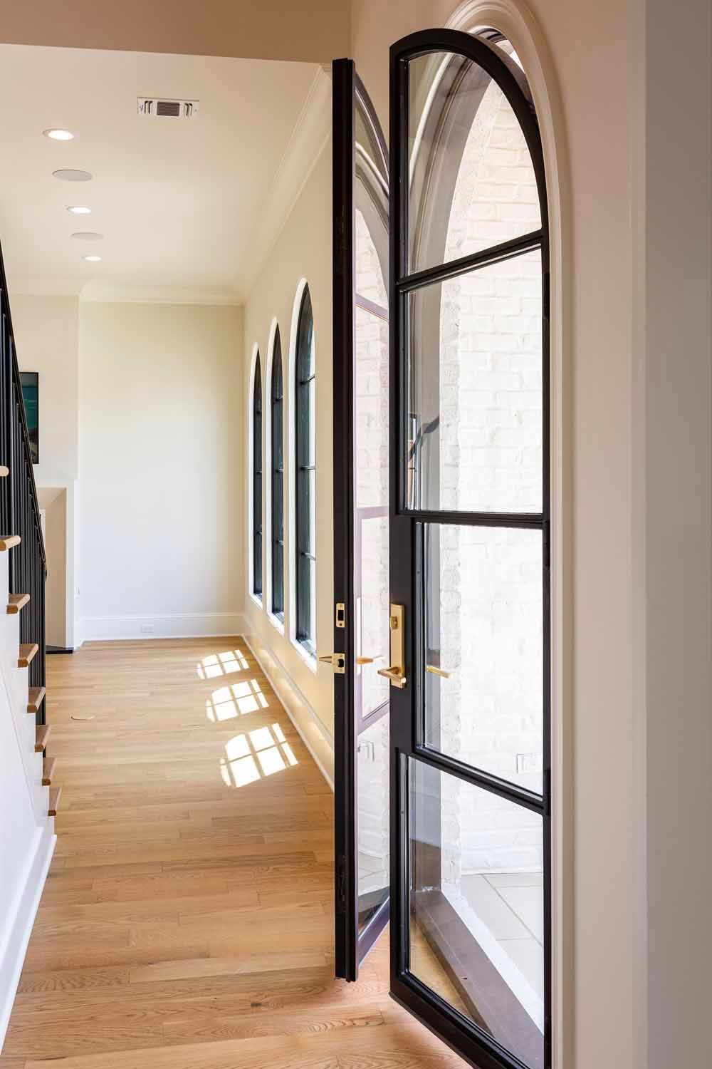 A view of a sunlit hallway with light wood floors and a row of black-framed arched glass doors and windows.