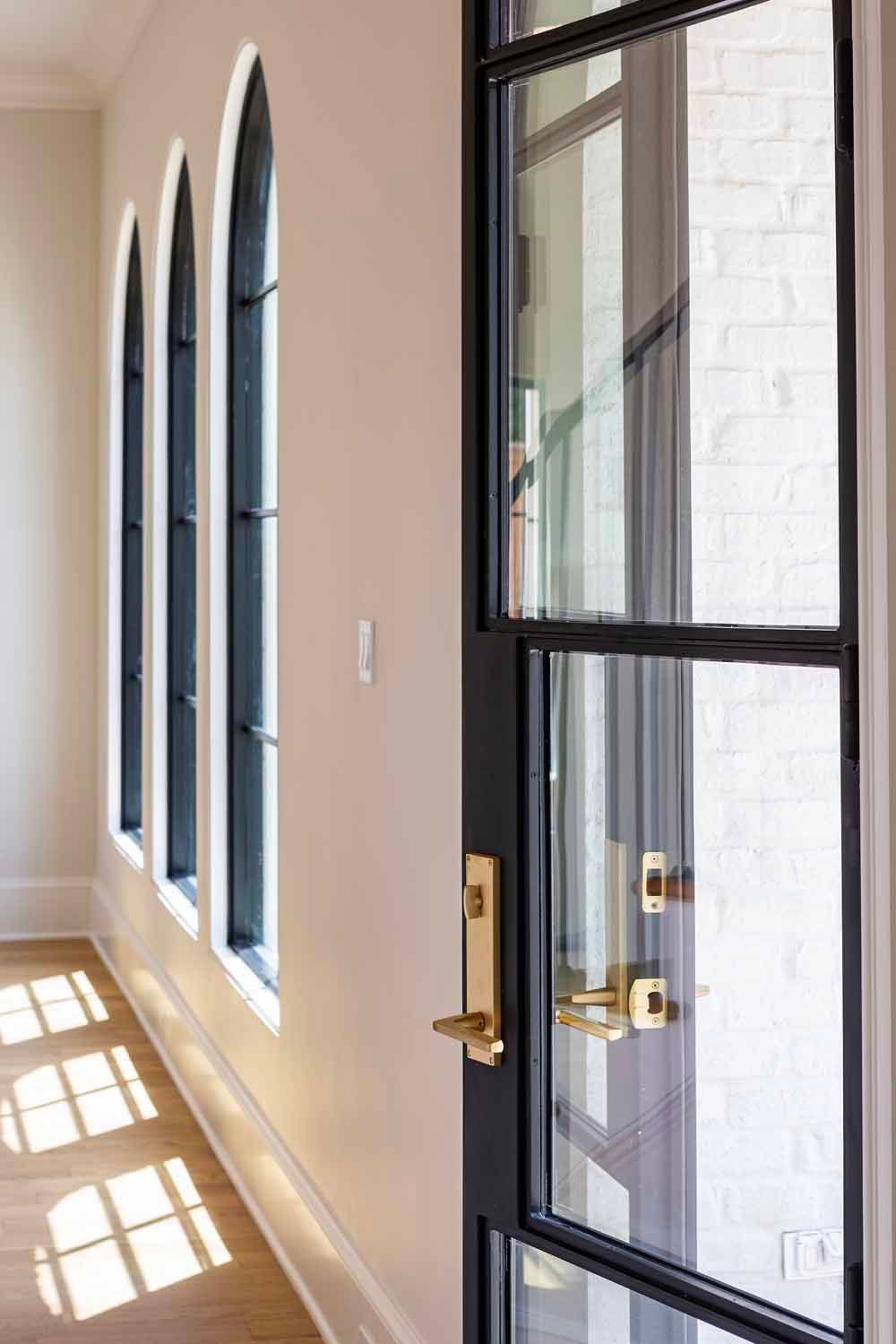 Black metal door with brass hardware next to a hallway with three arched windows and sunlight casting shadows on wood floors.