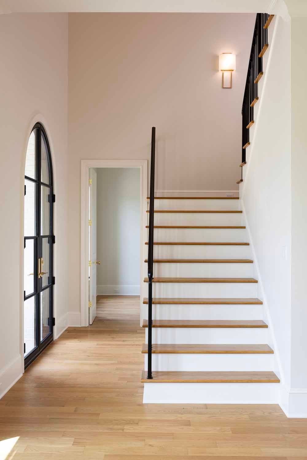 A bright foyer with a wood-floored staircase, black metal railings, an arched glass door, and a wall sconce.