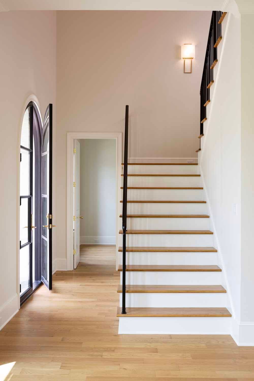 A bright foyer with light wood floors, a white staircase with a black handrail, an arched glass door, and a wall sconce.