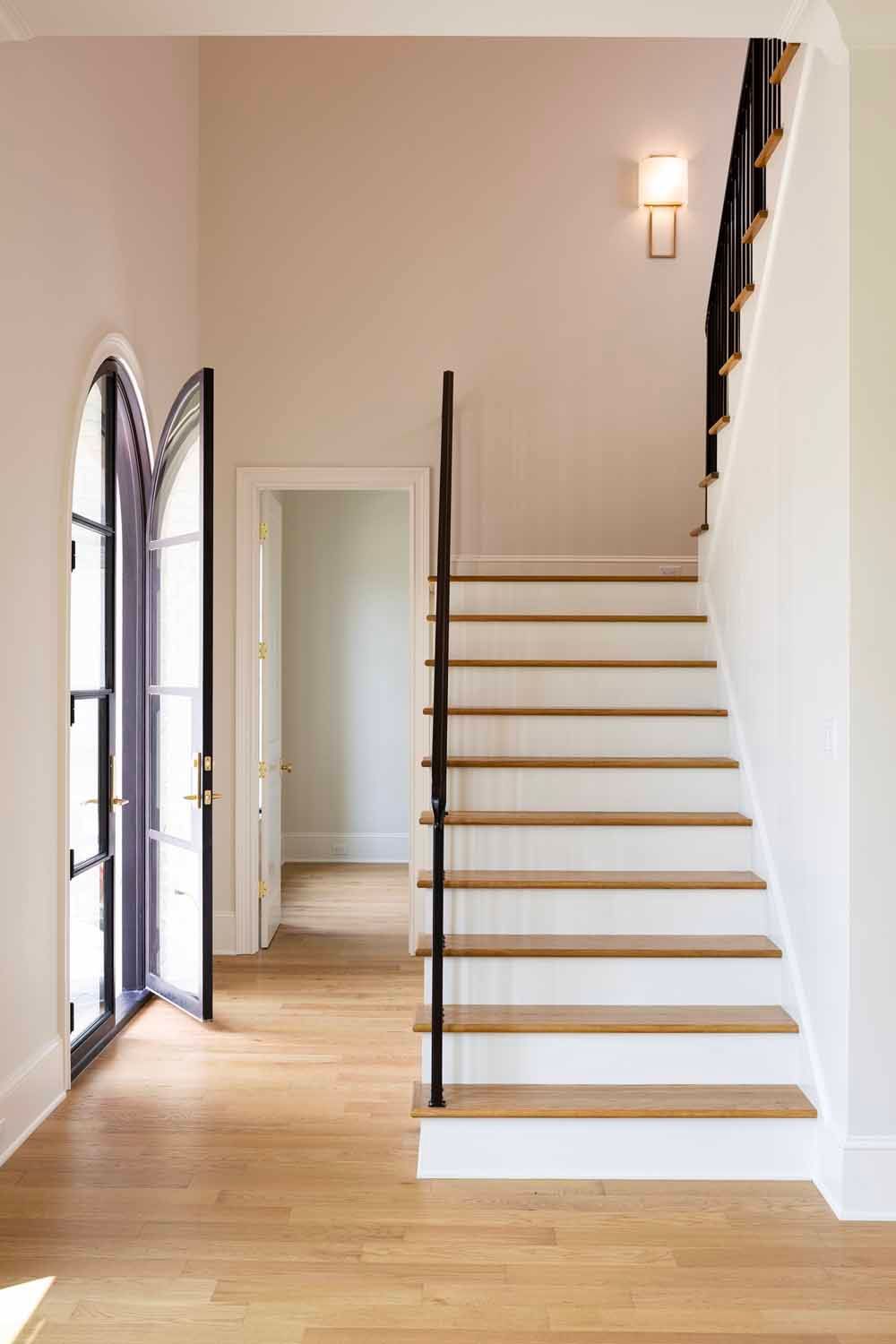 A bright foyer with a white staircase, light wood floors, an arched doorway, and a modern wall-mounted light fixture.