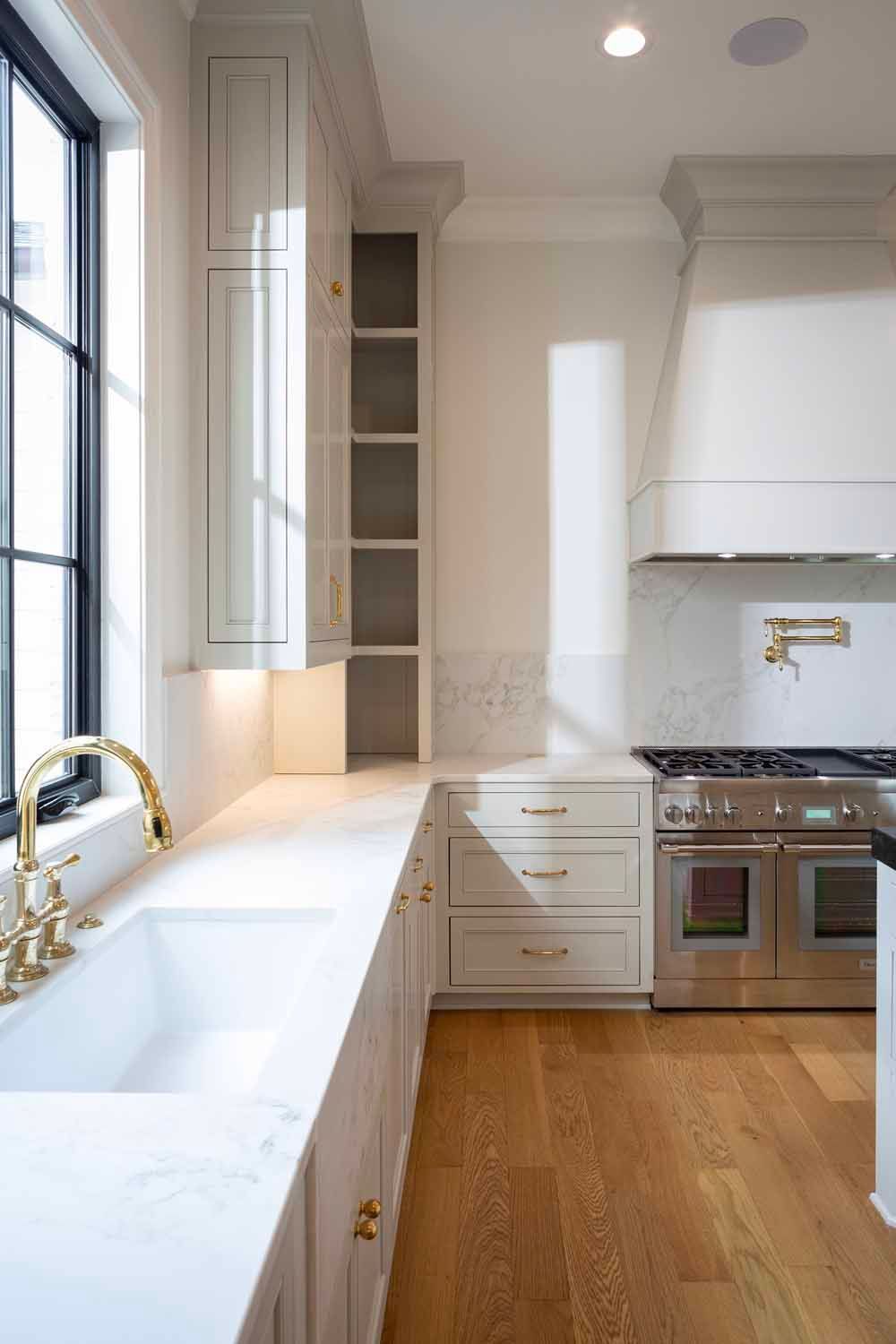 A modern kitchen featuring white cabinets, a farmhouse sink, gold faucet, marble countertops, and a professional range.