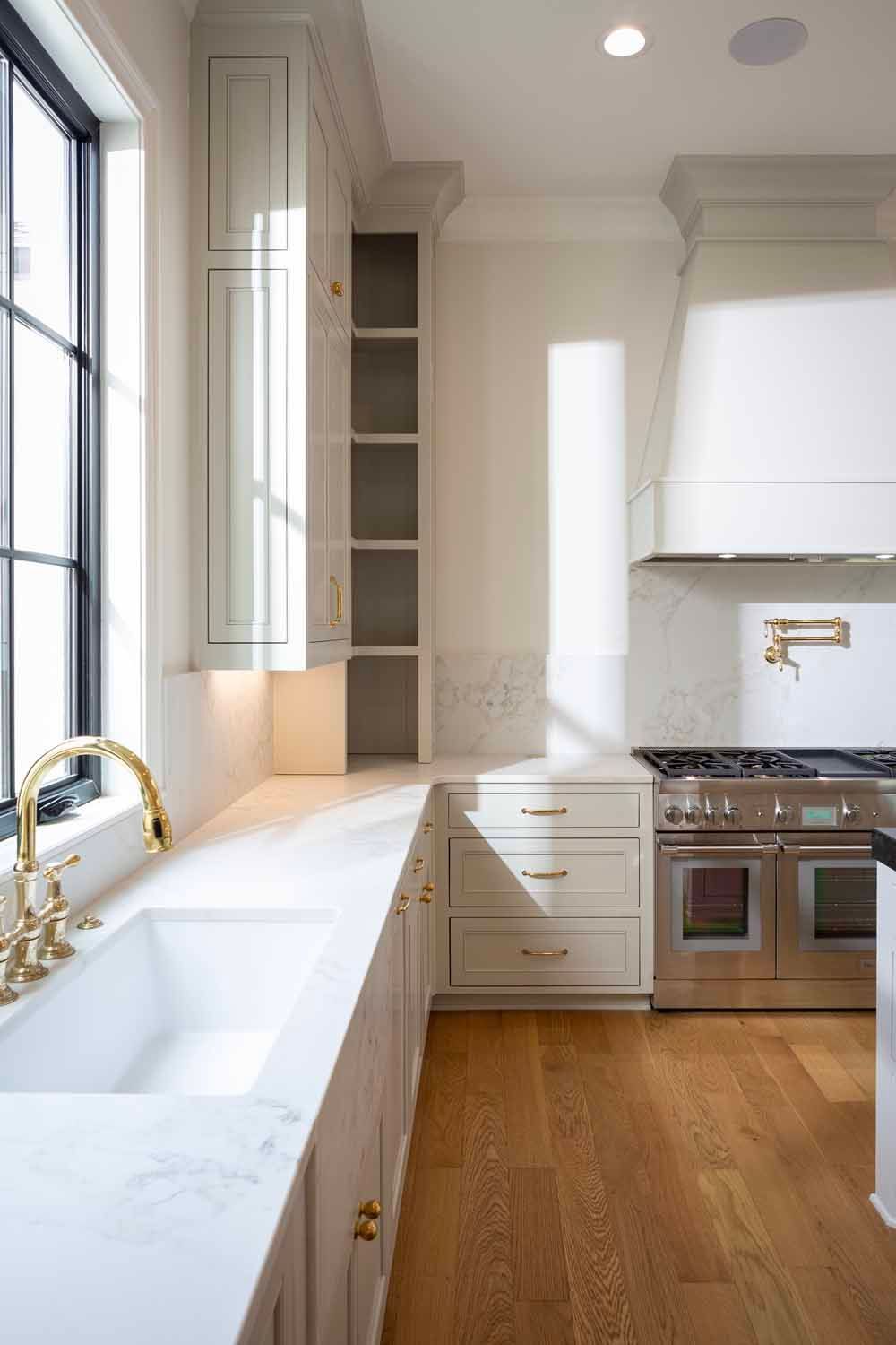 A bright kitchen with white marble counters, light grey cabinets, a farmhouse sink, gold fixtures, and a large stove.