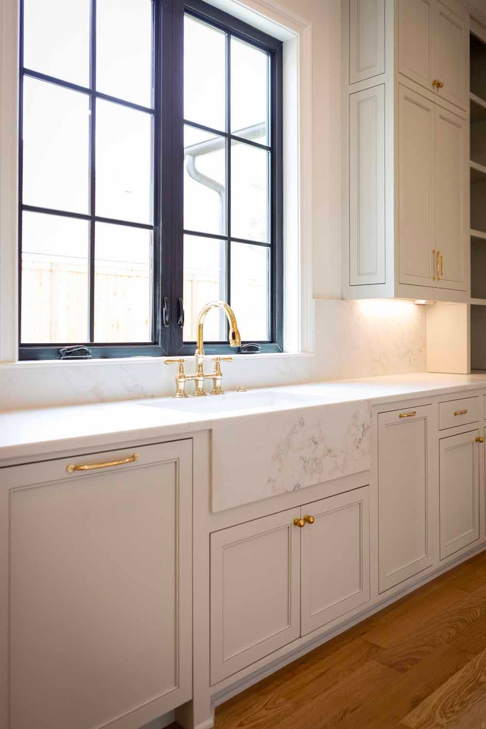 A bright, modern kitchen featuring white cabinets, a marble farmhouse sink, gold hardware, and a dark-framed window.