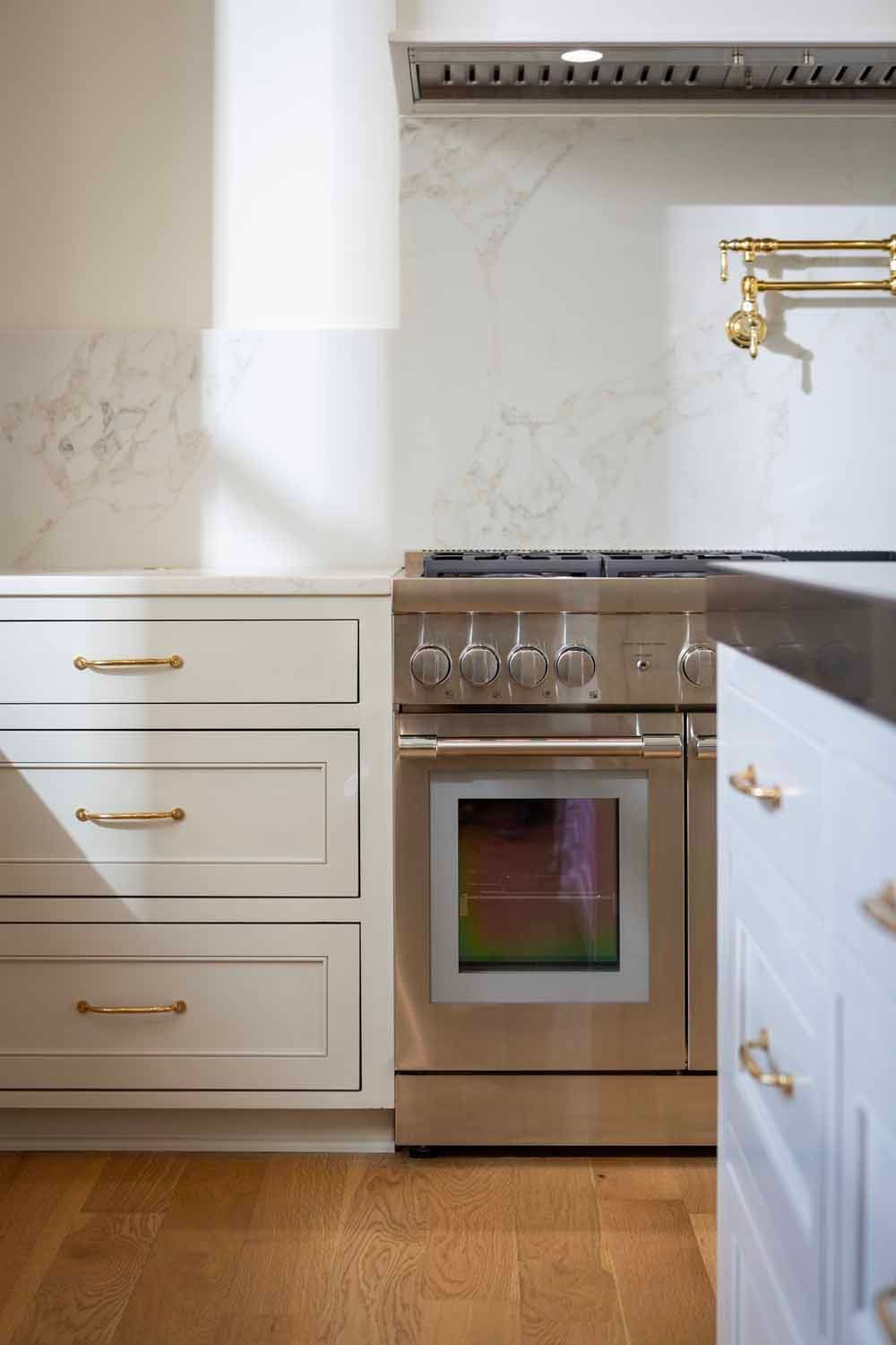 A modern kitchen with white drawers, a stainless steel range, marble backsplash, gold hardware, and light wood flooring.