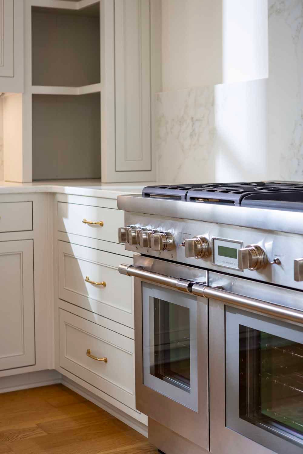 A professional stainless steel range sits in a bright kitchen with white cabinetry and wooden floors.