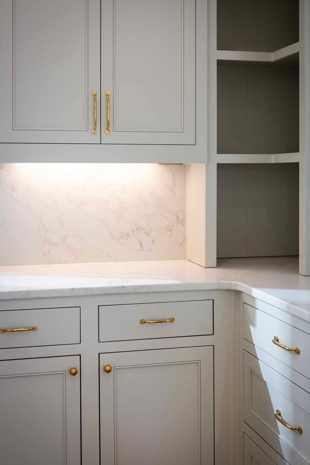 Light gray kitchen cabinets with gold handles and an illuminated marble backsplash, featuring open corner shelving.