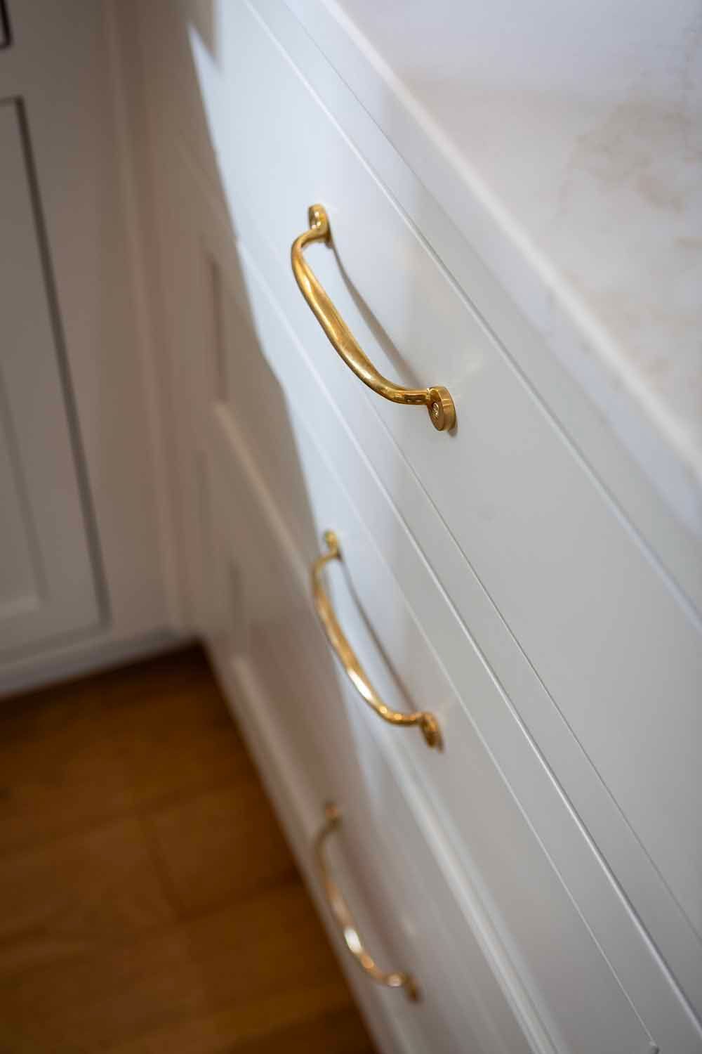 A close-up of three white kitchen drawers with ornate gold handles, set against a wooden floor.
