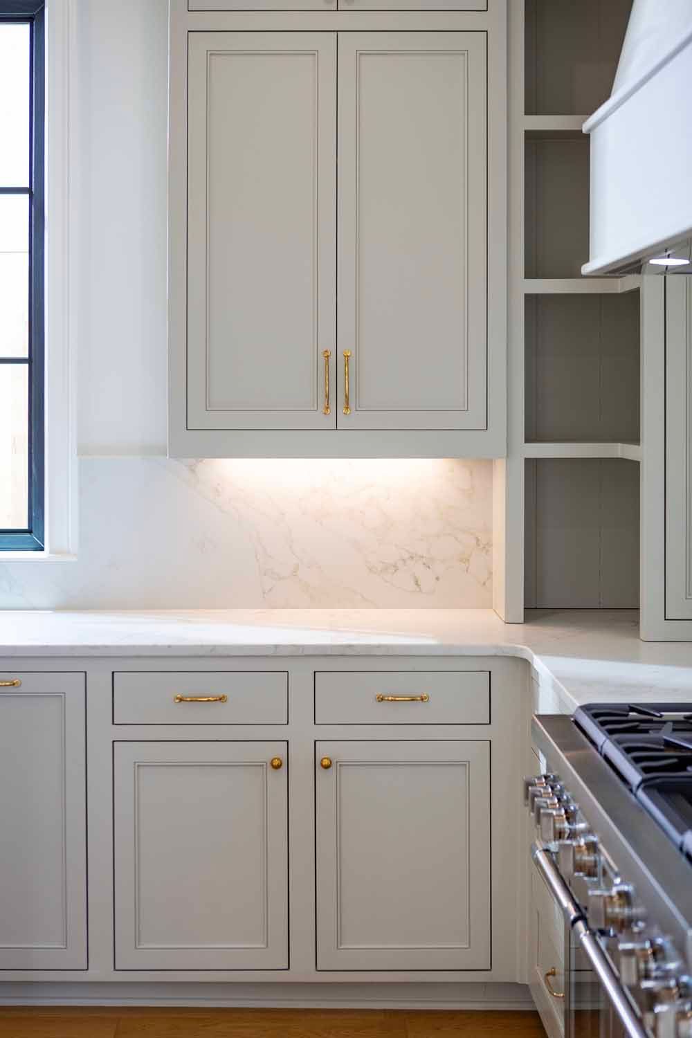 Light gray kitchen cabinetry and a marble countertop with under-cabinet lighting and a stainless steel stove.