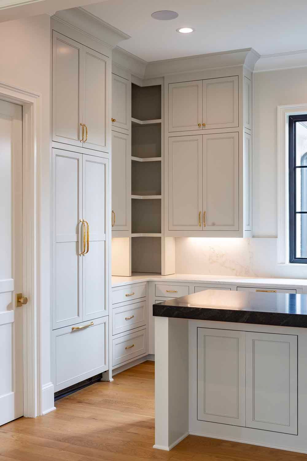 A kitchen corner with light grey cabinets, gold hardware, open corner shelving, and a dark countertop on a kitchen island.