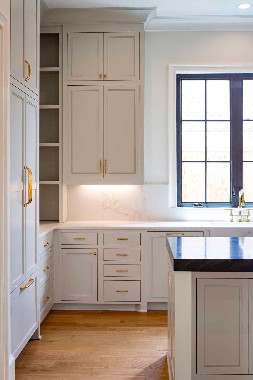 A modern kitchen featuring light grey cabinets, brass hardware, wood flooring, and a large window with a dark frame.