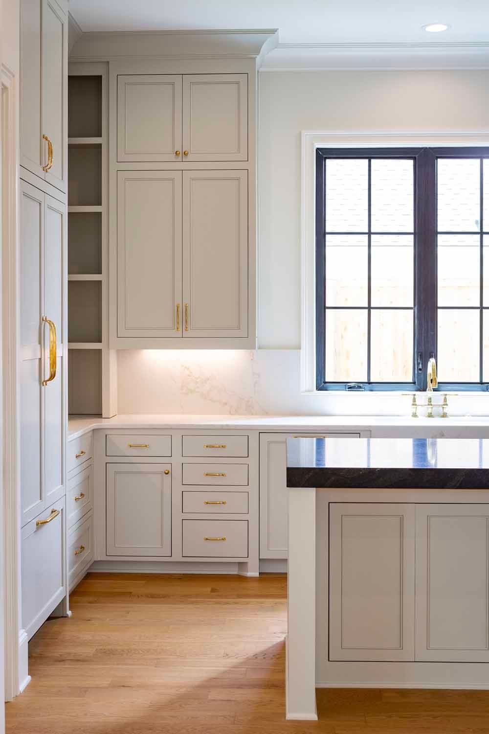 A bright, modern kitchen features light gray cabinets, gold hardware, a marble backsplash, and a dark island countertop.