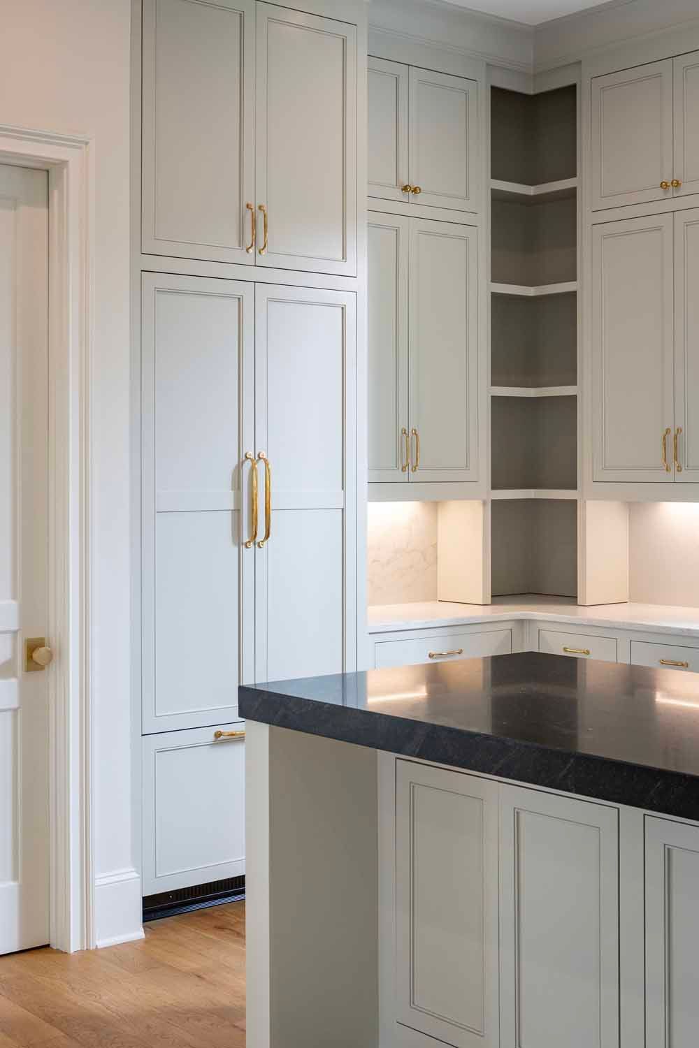 Light gray kitchen cabinets with gold hardware, open shelving, and a dark stone countertop in a bright kitchen.