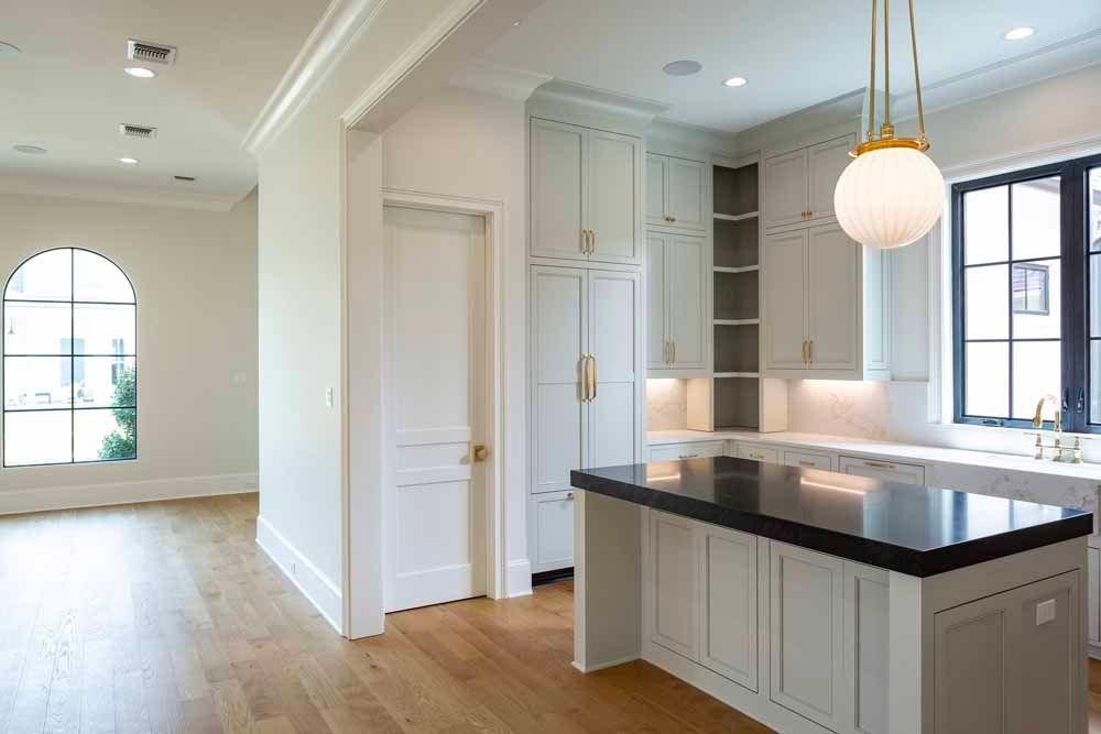 A modern kitchen with a dark countertop island, light grey cabinets, and a hanging pendant light next to an arched window.