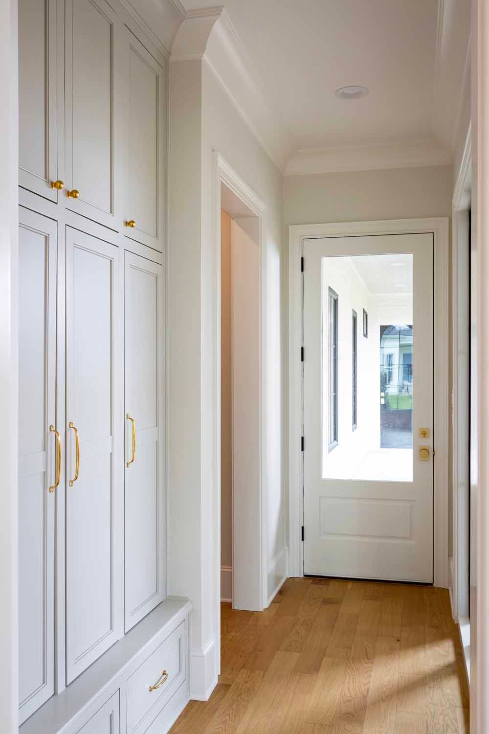 Light gray built-in cabinets with gold hardware line a bright hallway with light wood floors and a glass-paned door.