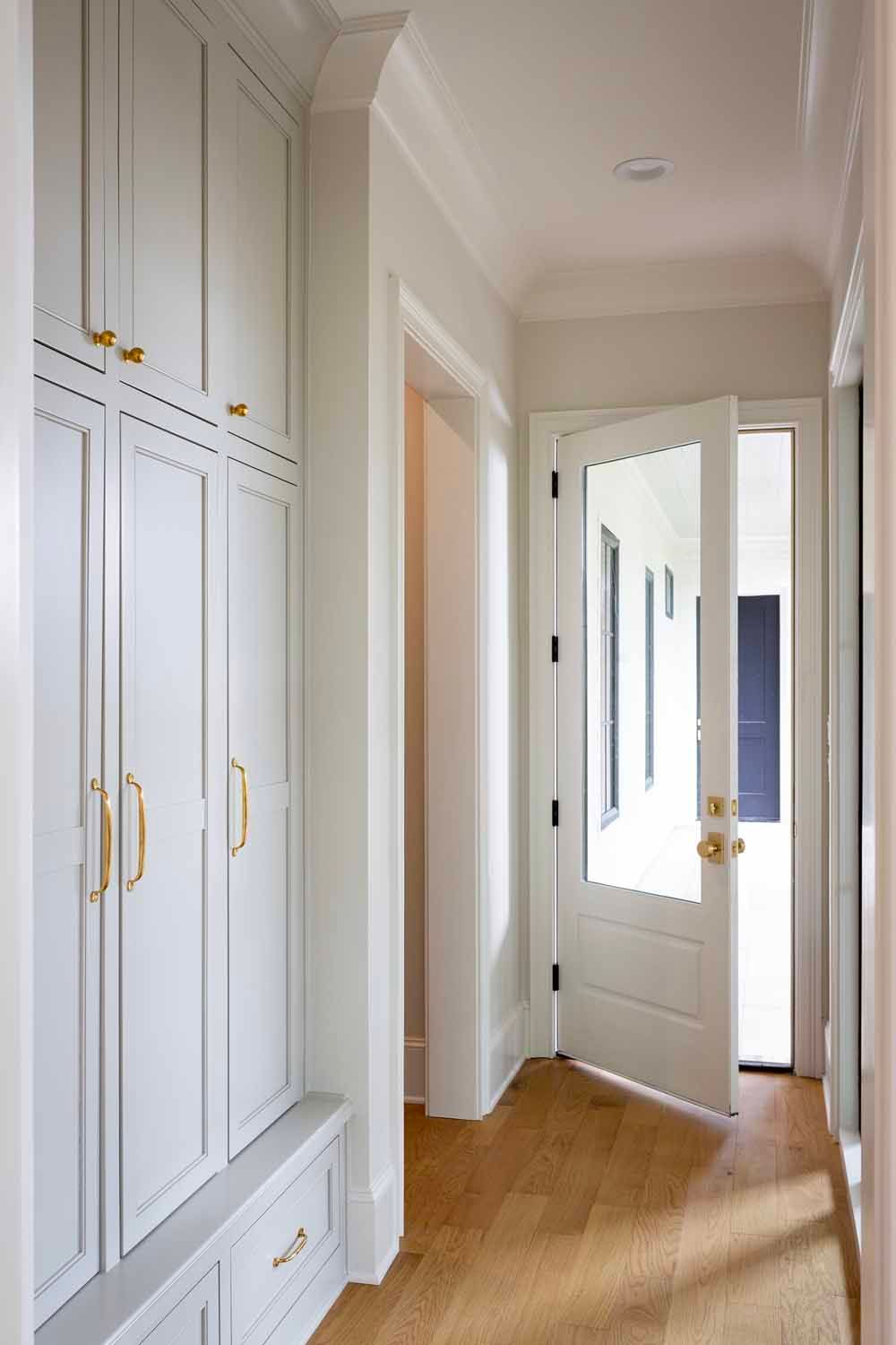 A hallway with light gray built-in cabinetry, brass hardware, wood floors, and an open glass-paned door leading outside.