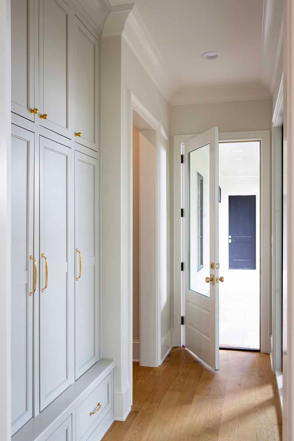 A hallway with light gray built-in cabinets and a bench, leading to an open door revealing an outdoor view.
