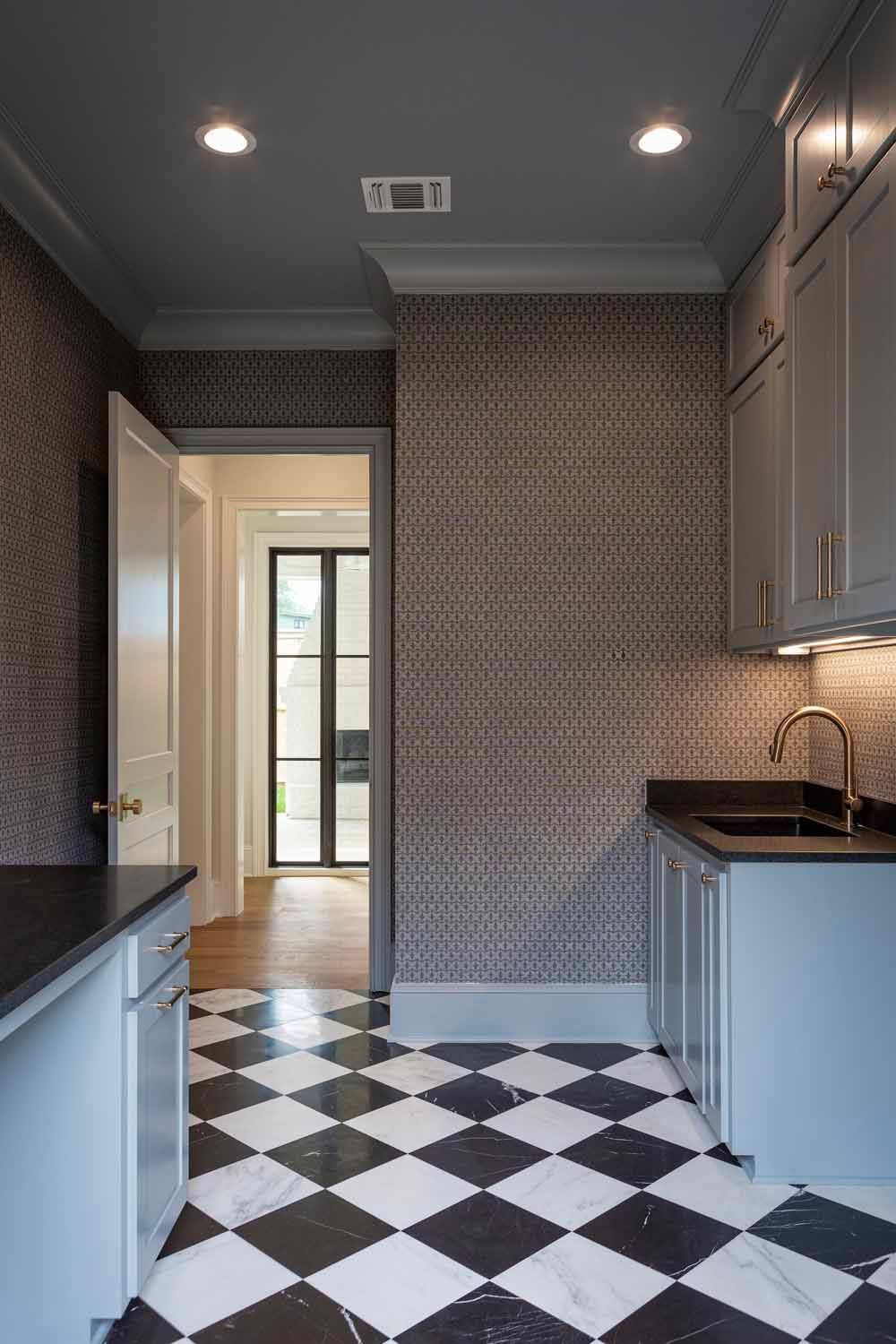 A butler's pantry with black and white checkered marble floors, gray cabinetry, dark countertops, and patterned wallpaper.