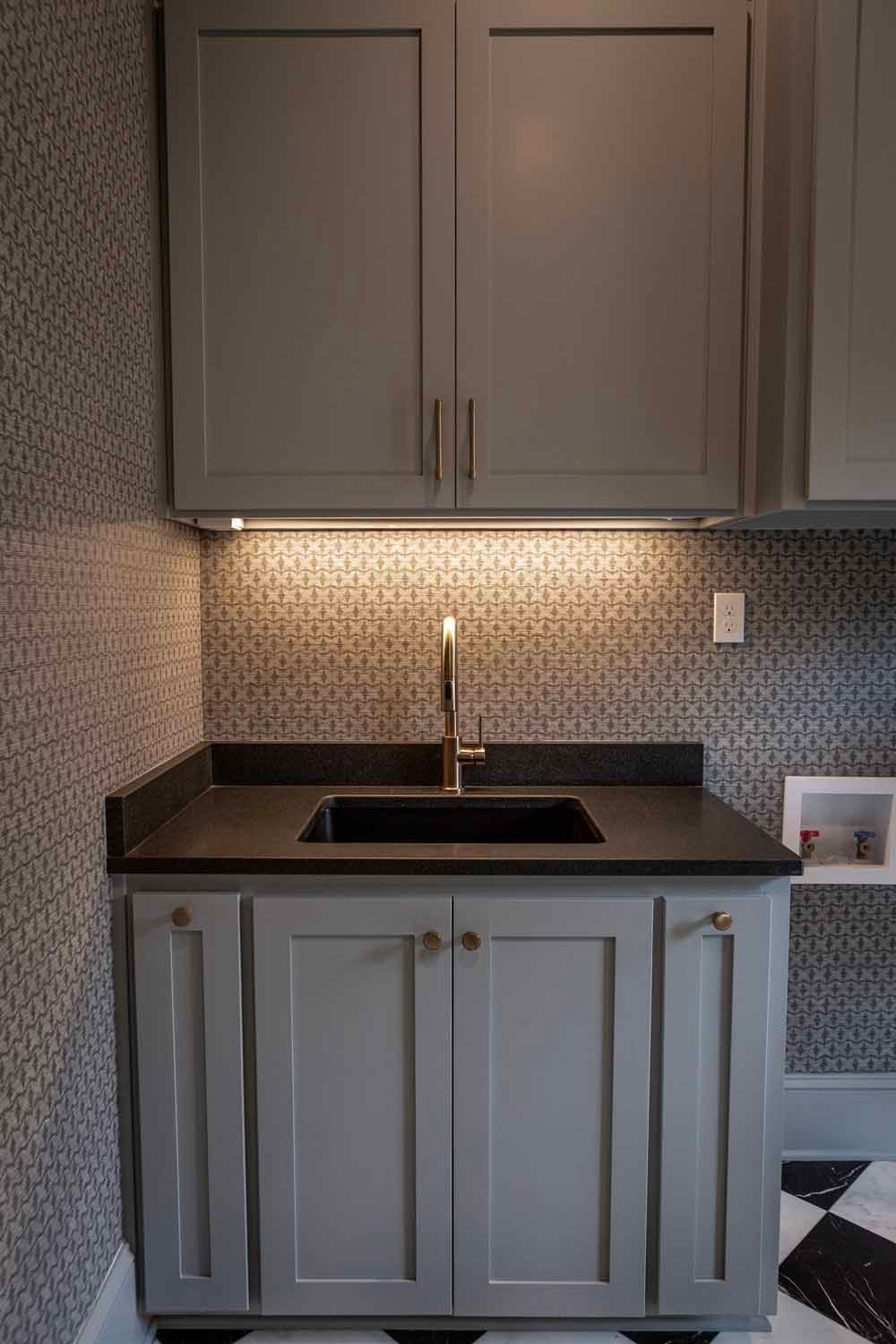 A kitchen wet bar with grey cabinets, a dark countertop, a small sink, and patterned wallpaper under under-cabinet lighting.