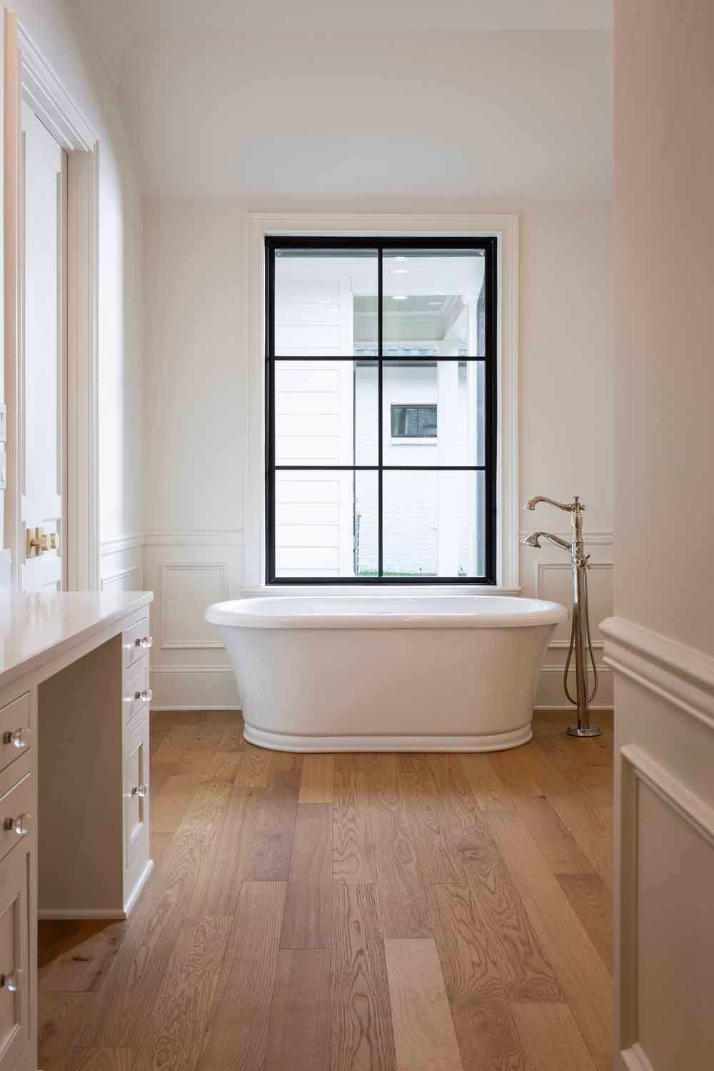 A white freestanding bathtub sits in front of a window with black grid frames in a room with light wood floors.