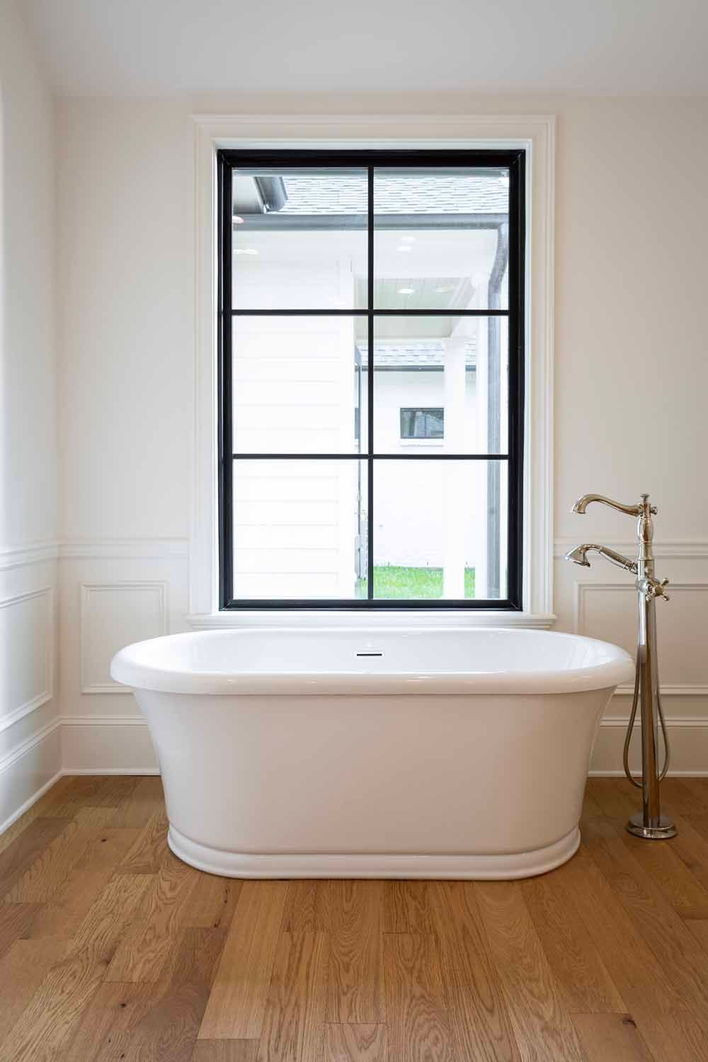 A white freestanding bathtub sits on a light wood floor in front of a window with a black frame and grid pattern.
