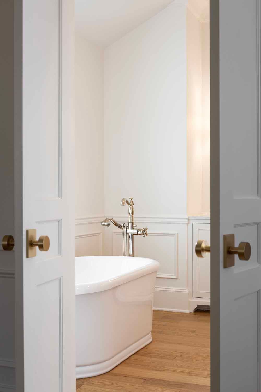 A view through white double doors into a bright bathroom featuring a freestanding white tub and brass fixtures.
