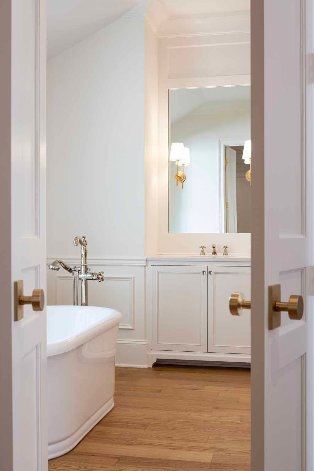 A view through a white door into a bright bathroom featuring a white soaking tub, wood floors, and a vanity with a mirror.