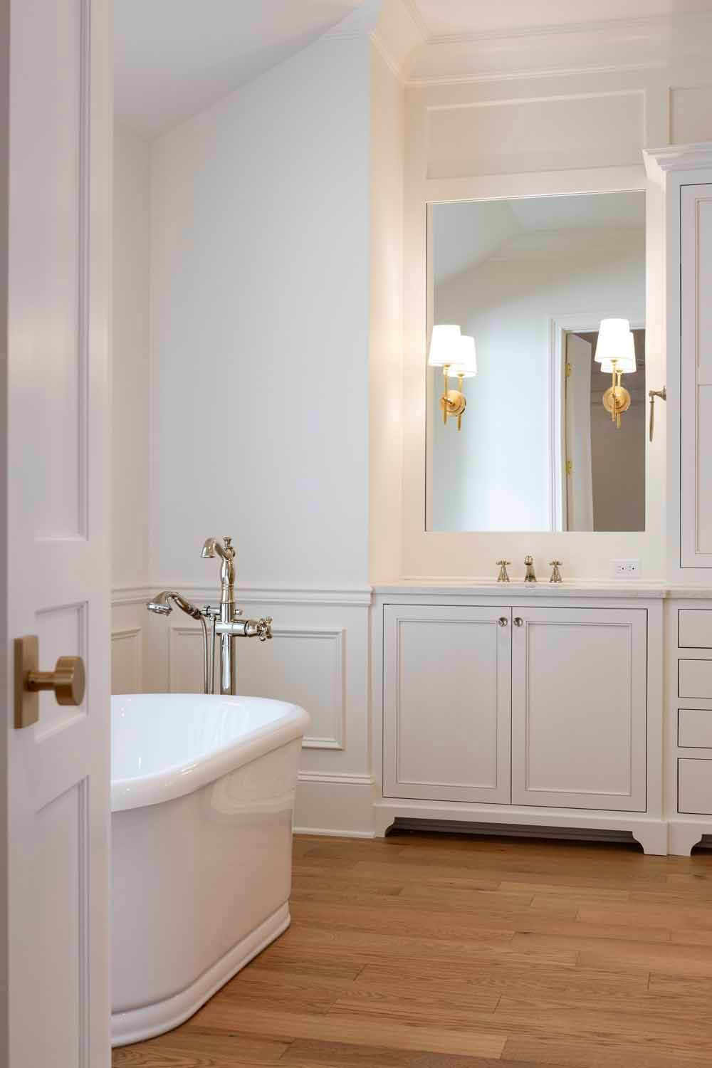 A white freestanding bathtub and vanity in a bright, modern bathroom with white cabinetry and light wood floors.