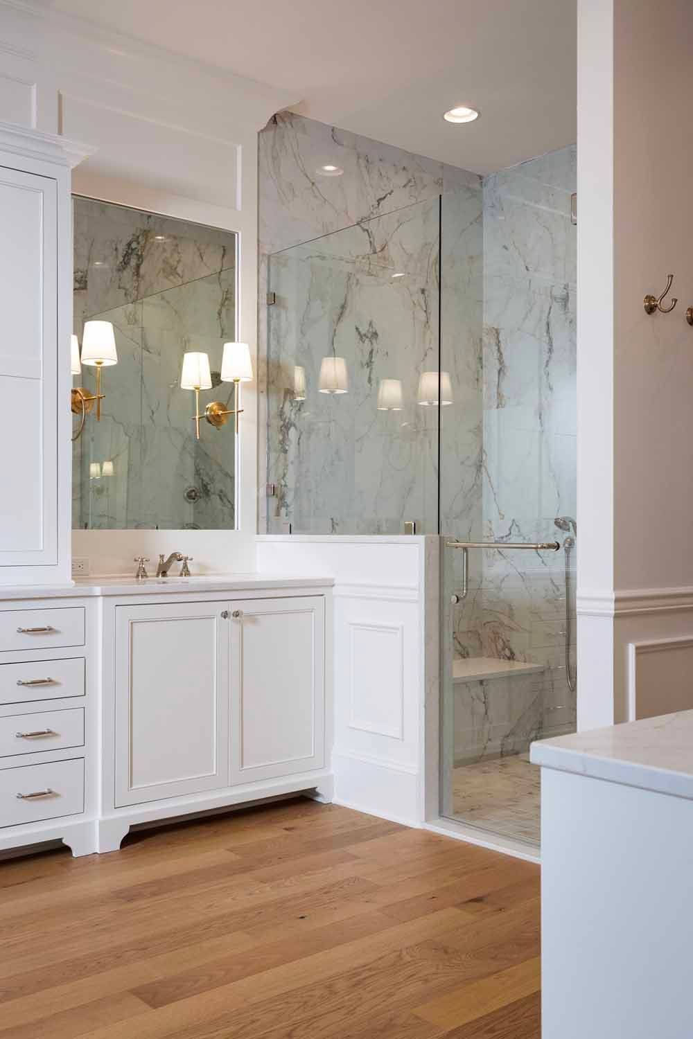 A modern bathroom with a white vanity, marble shower wall, glass enclosure, and light wood flooring.
