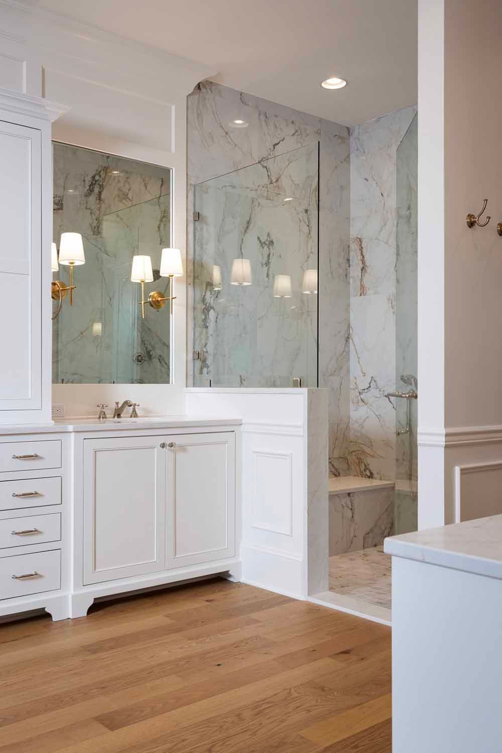 A bright bathroom with white vanity cabinets, light wood flooring, marble-tiled walk-in shower, and brass wall sconces.