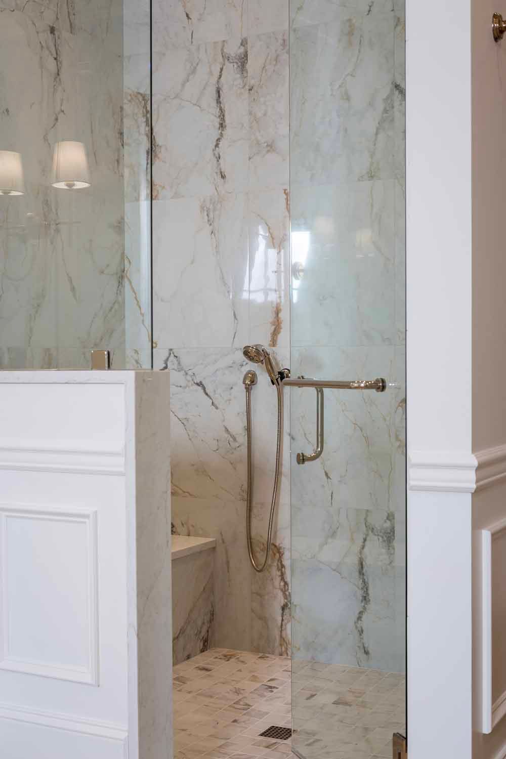 A glass shower stall with marble walls and a metallic handheld showerhead inside a bathroom with white paneled walls.