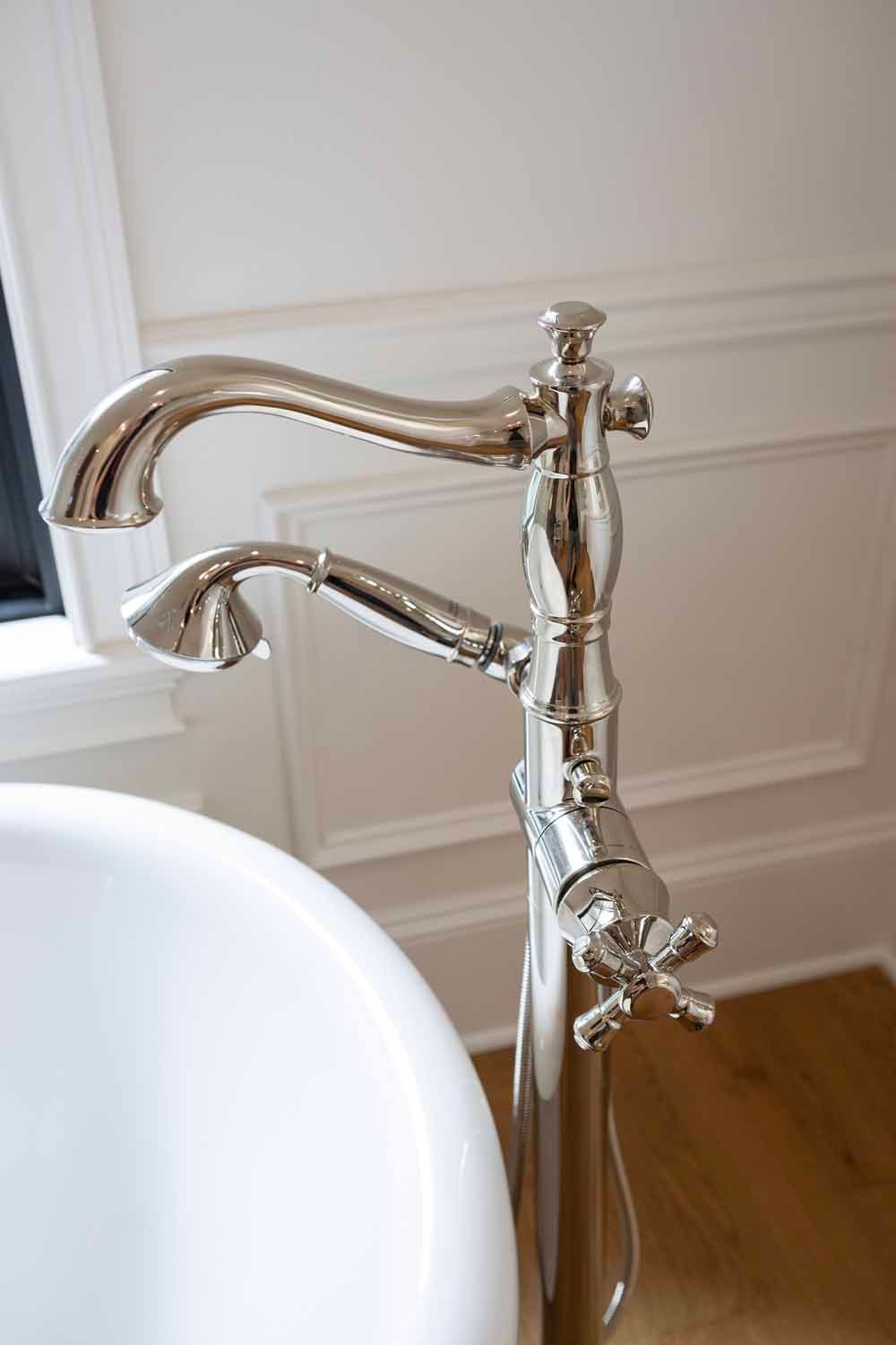 A chrome floor-mounted bathtub faucet with a hand shower attachment, positioned next to a white tub against a paneled wall.