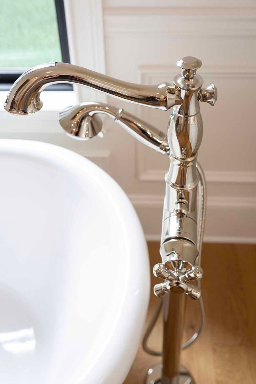 A polished chrome, floor-mounted bathtub faucet with a hand shower attachment next to a white porcelain tub.