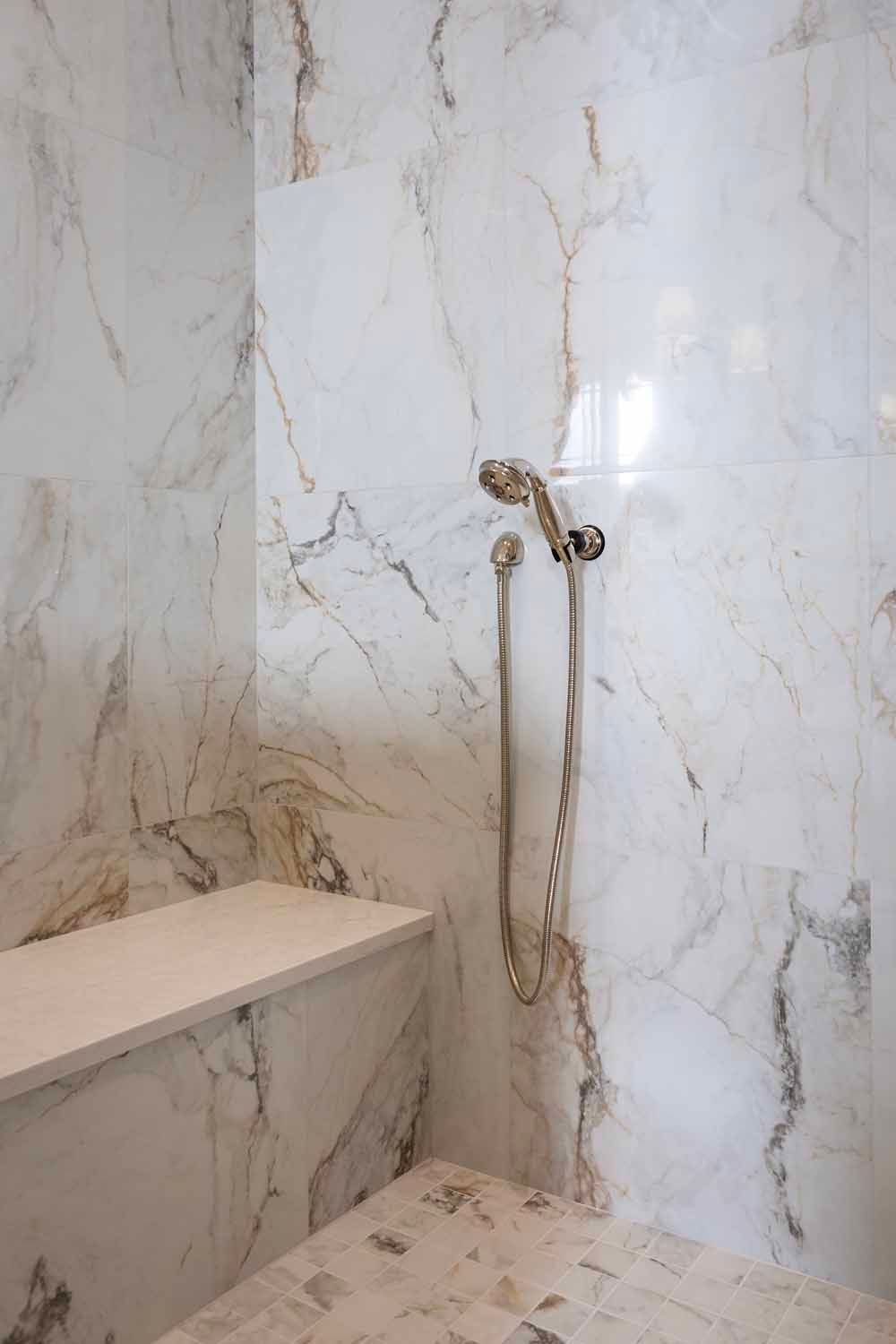 A shower area with white and gold marble walls, a built-in bench, and a polished nickel handheld shower head.