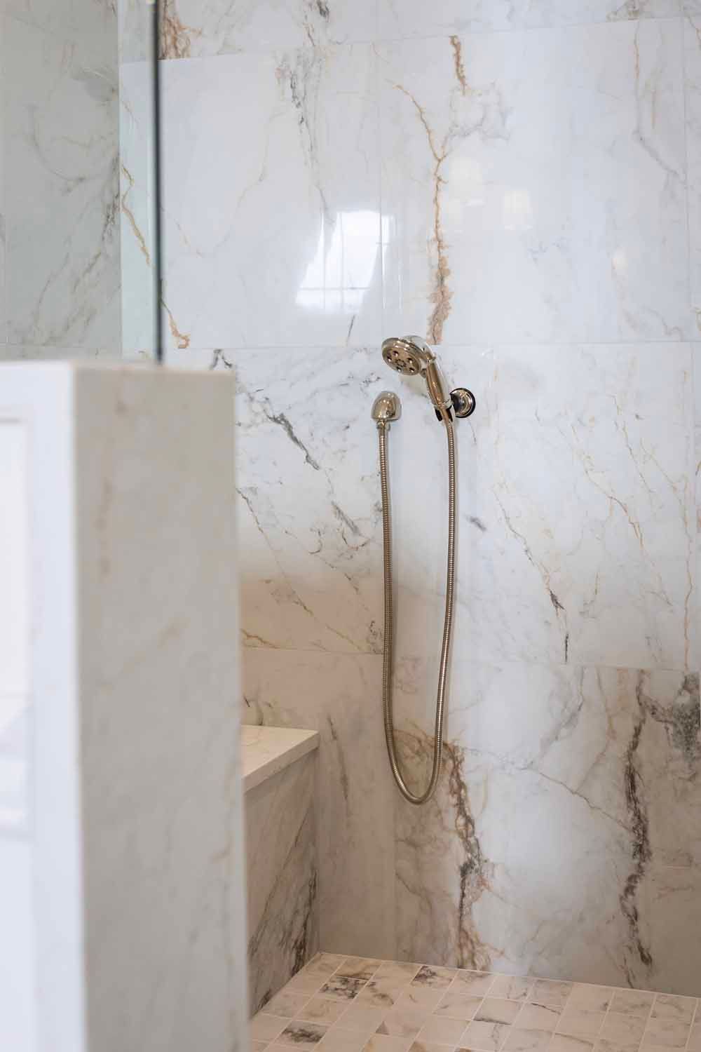 A gold-finished handheld shower fixture mounted on white marble tiled walls.