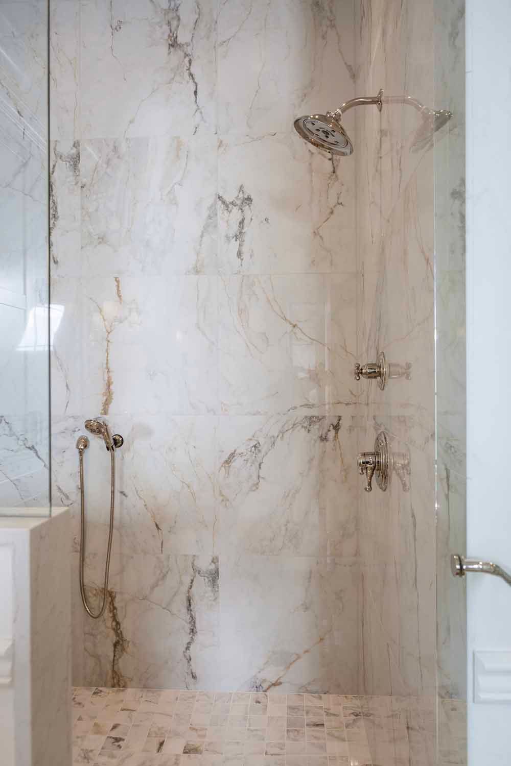 Modern shower featuring light marble wall tiles, metallic fixtures, a handheld wand, and a glass door enclosure.