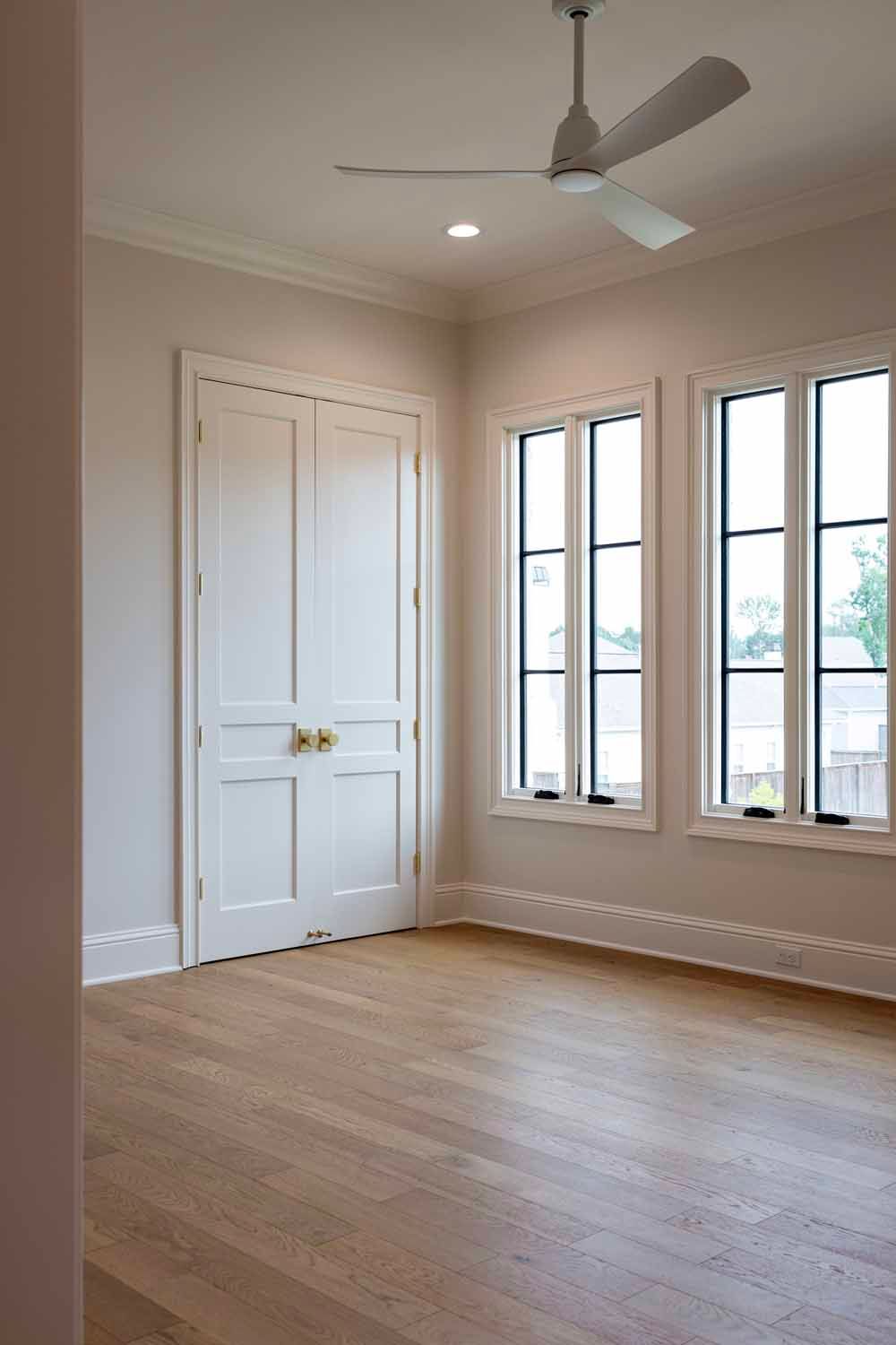 An empty room with light wood floors, neutral walls, a white double door with brass knobs, and two windows with black frames.