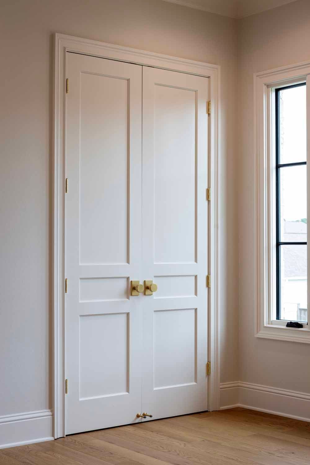 White double doors with brass knobs and hinges, set in a light-colored wall next to a window with a black frame.