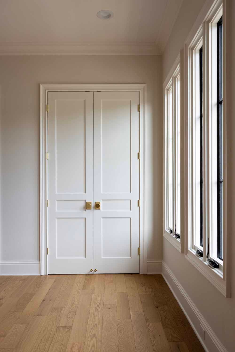 White double doors with brass knobs and nearby windows in a room with hardwood floors and light-colored walls.